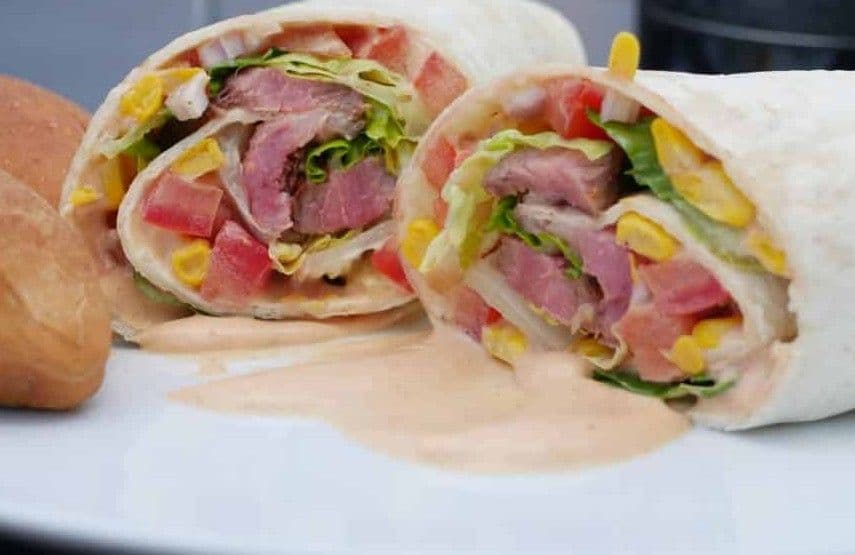 Angeschnittener Steak Burrito mit Entrecote-Streifen, Mais, Tomate und Salat, daneben Brötchen und Sauce