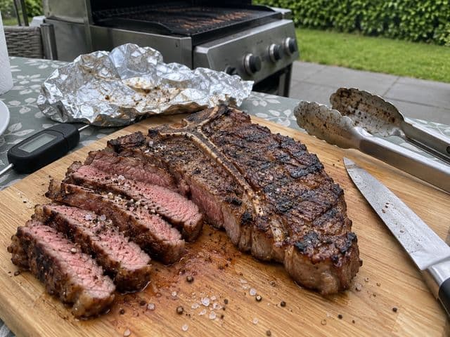 Gegrilltes T-Bone-Steak auf dem Holzbrett, die Roastbeef-Seite in Scheiben geschnitten mit rosa Kern, daneben Thermometer und Alufolie, im Hintergrund der Gasgrill