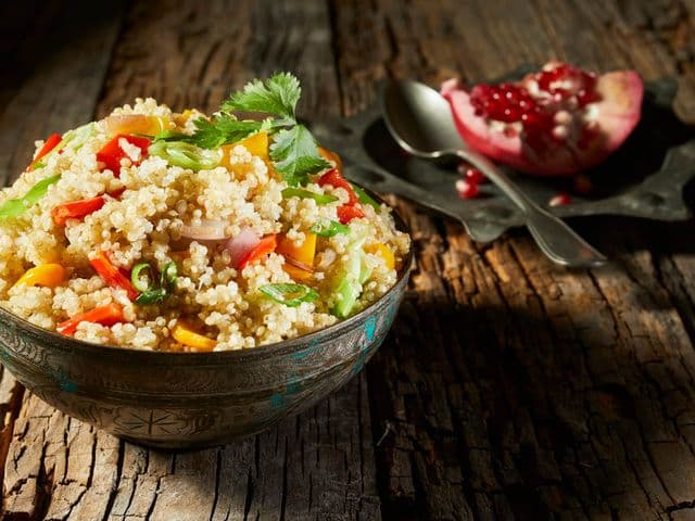 Hier liegt eine Schale mit Couscous Salat und im Hintergrund ein Granatapfel mit einem Löffel auf einem Teller.