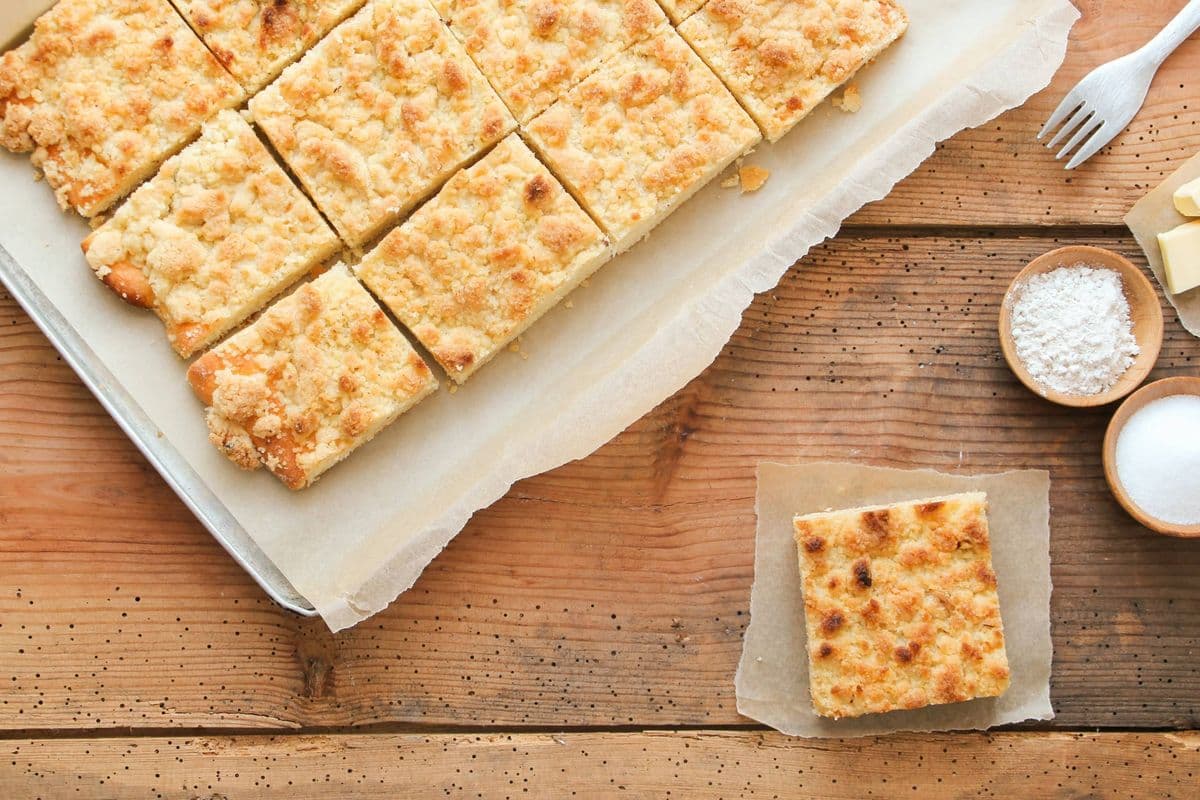 Blech mit mehreren Stücken Streuselkuchen auf einem Holztisch. Daneben eine Plastikgabel und die Zutaten für das Rezept.