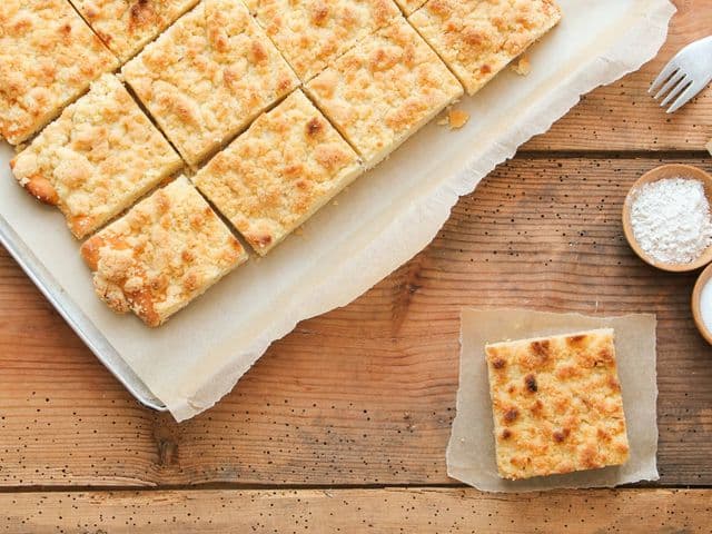 Blech mit mehreren Stücken Streuselkuchen auf einem Holztisch. Daneben eine Plastikgabel und die Zutaten für das Rezept.
