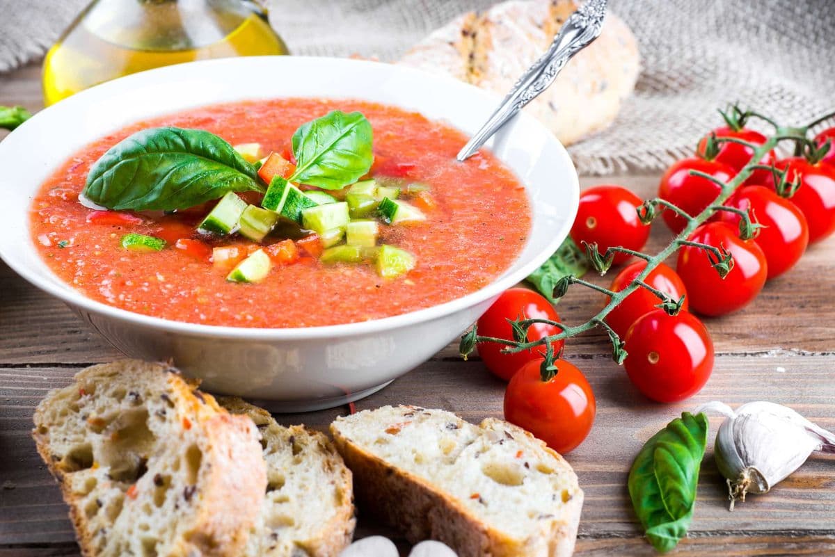 Eine Schüssel mit Gazpacho-Suppe und daneben Cherrytomaten, eine Knobauchzehe, etwas Basilikum, Öl und frisches Brot.