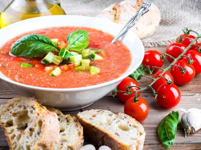 Eine Schüssel mit Gazpacho-Suppe und daneben Cherrytomaten, eine Knobauchzehe, etwas Basilikum, Öl und frisches Brot.