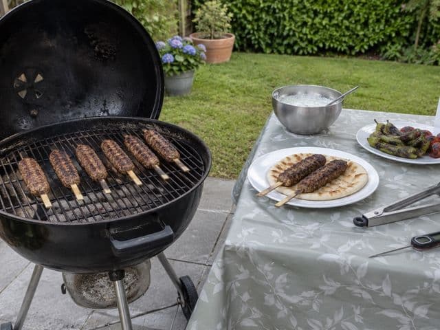 Köfte-Spieße auf dem Kugelgrill im Garten, daneben Fladenbrot mit Köfte, Joghurtsauce und Grillgemüse auf dem Gartentisch