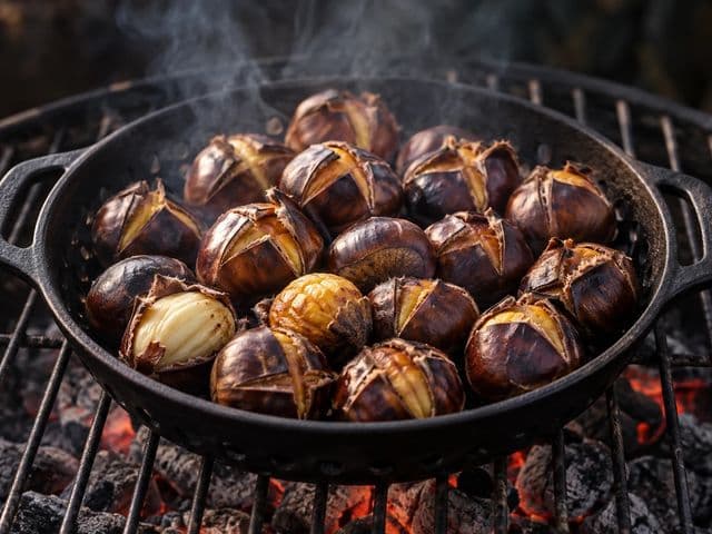 Gegrillte Maronen in einer Gusseisen-Grillschale auf dem Rost, die kreuzförmig eingeschnittenen Schalen haben sich aufgerollt, einzelne Maronen halb geschält