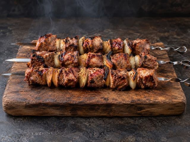 Russisches Schaschlik: drei Metallspieße mit gegrillten Schweinefleisch- und Zwiebelwürfeln auf einem Holzbrett, ohne Sauce