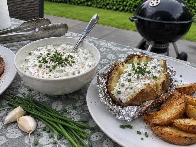 Selbstgemachte Sour Cream mit Schnittlauch in einer Schüssel, daneben eine Folienkartoffel mit Sour Cream, Kartoffel-Wedges und Steak auf dem Gartentisch
