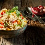 Hier liegt eine Schale mit Couscous Salat und im Hintergrund ein Granatapfel mit einem Löffel auf einem Teller.