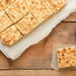 Blech mit mehreren Stücken Streuselkuchen auf einem Holztisch. Daneben eine Plastikgabel und die Zutaten für das Rezept.